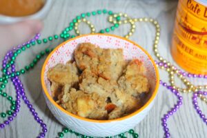 Cafe au Lait Bread Pudding in a white bowl with an orange rim surrounded by mardi gras beads with a Cafe du Monde coffee can in the background