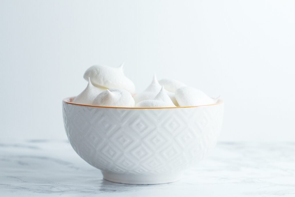 White textured bowl with orange rim filled with white meringues in front of a white background on a marbled surface