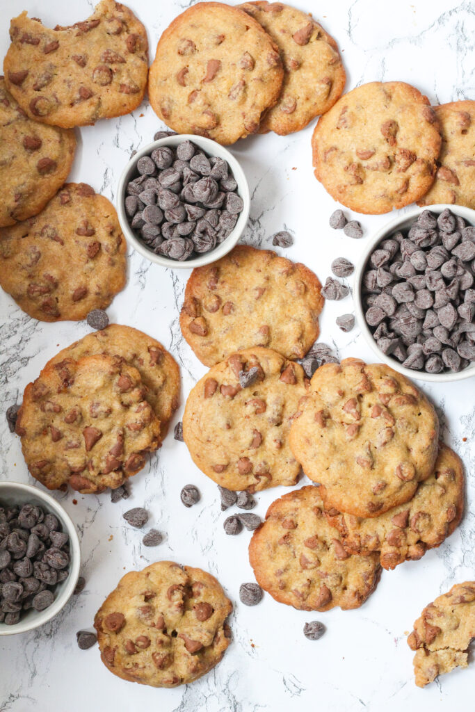 Top down view of a lot of cookies with three small bowls of chocolate chips and loose chocolate chips  spread around throughout the cookies