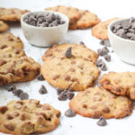 Angled view of a lot of cookies with three small bowls of chocolate chips and loose chocolate chips spread around throughout the cookies