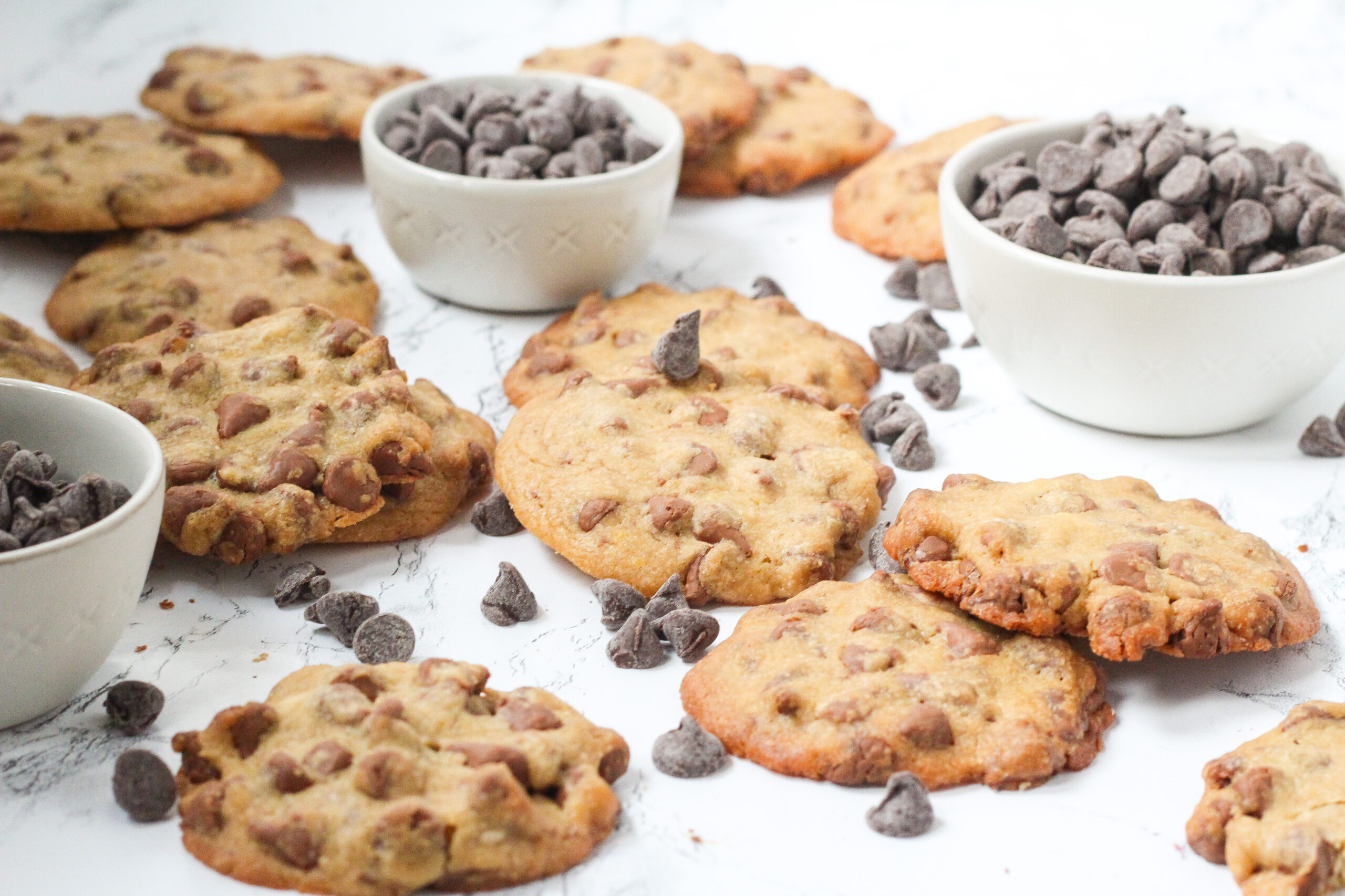 Angled view of a lot of cookies with three small bowls of chocolate chips and loose chocolate chips spread around throughout the cookies
