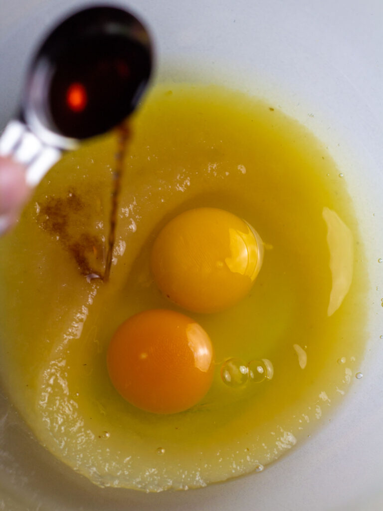 A measuring spoon of vanilla being poured into a mixing bowl with applesauce and eggs.