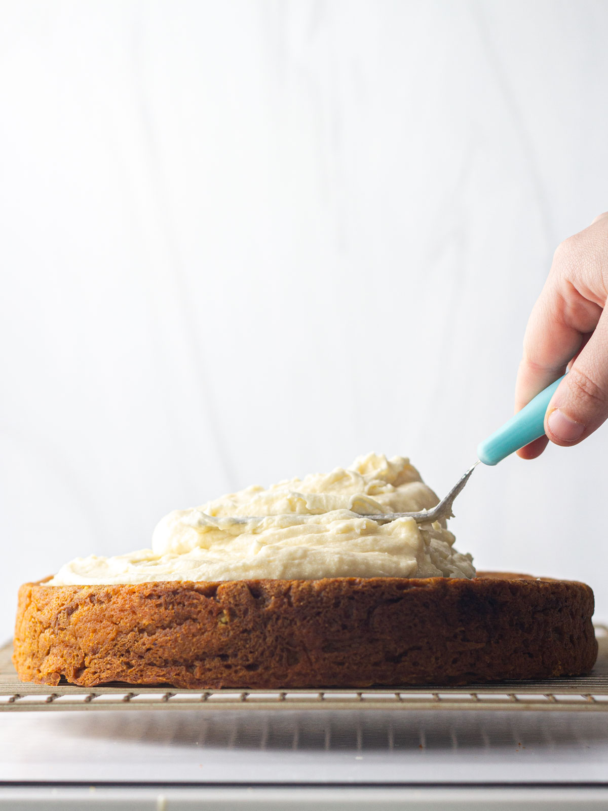 A hand spreading marshmallow frosting onto a sweet potato cake on a gold wire rack.