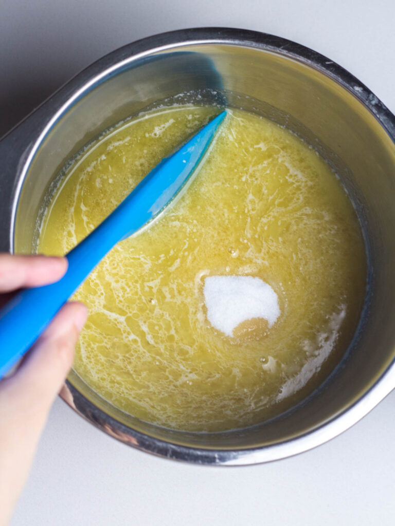 A hand holding a silicones spatula stirring sugar into the melted butter and apple cider mixture.