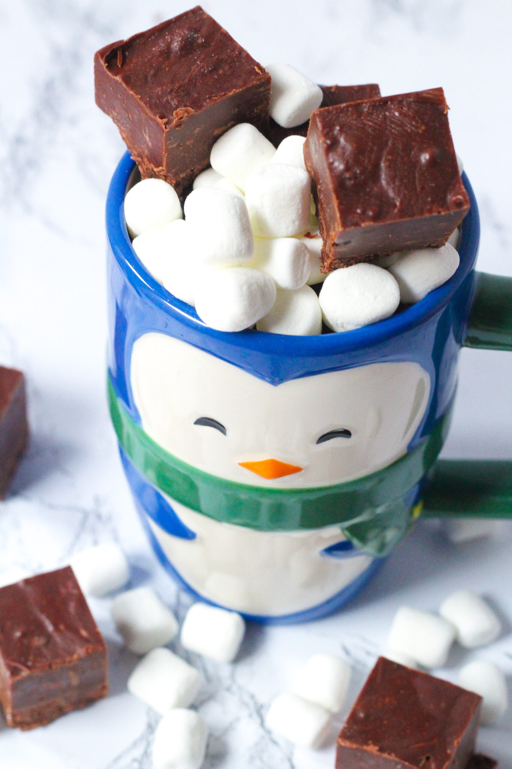 angled top down view of a really cute penguin mug filled with mini marshmallows and two squares of brown hot cocoa fudge on top, and with mini marshmallows and fudge surrounding the mug.