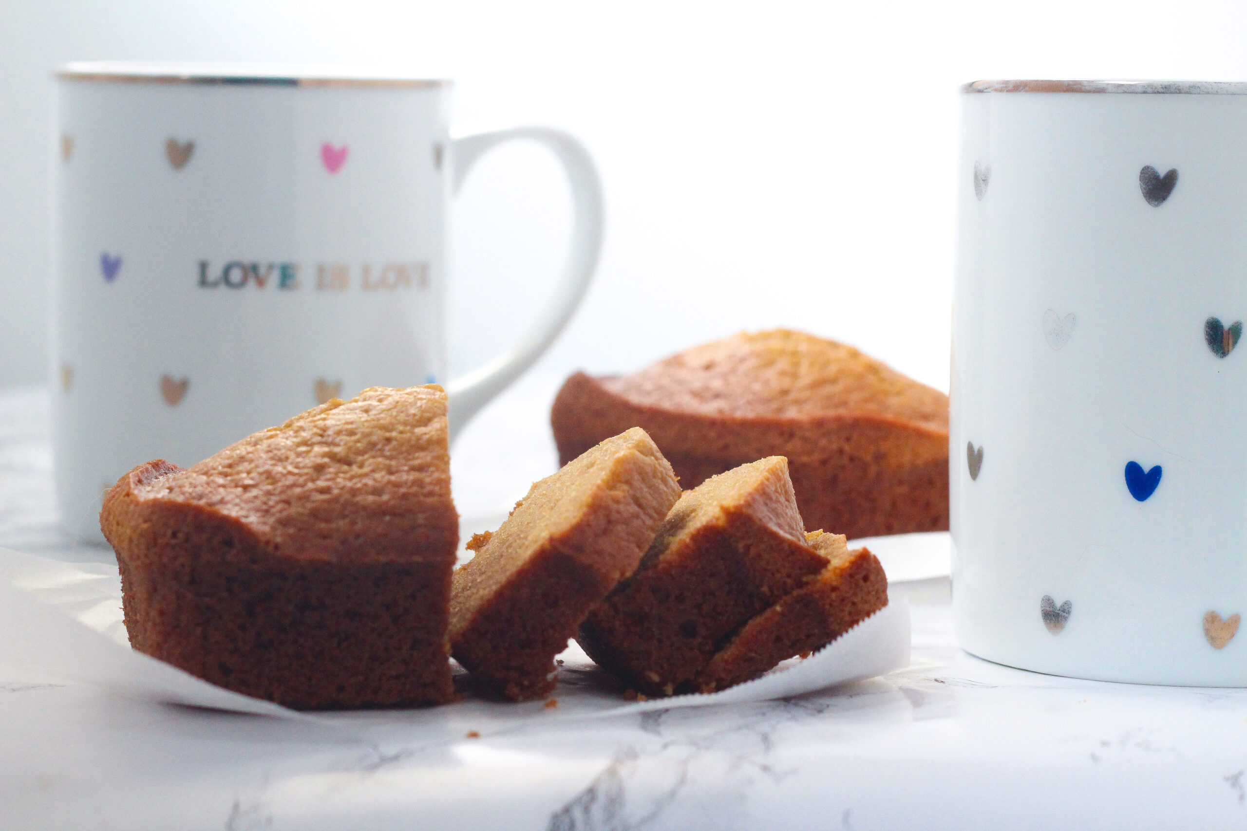 Side view of a partially sliced chai loaf with a whole chai loaf in the background. Behind the sliced chai loaf toward the left is a white mug with colorful hearts that says love is love, and next to the sliced chai loaf to the right, cut off in the frame, is a matching mug, but without the words