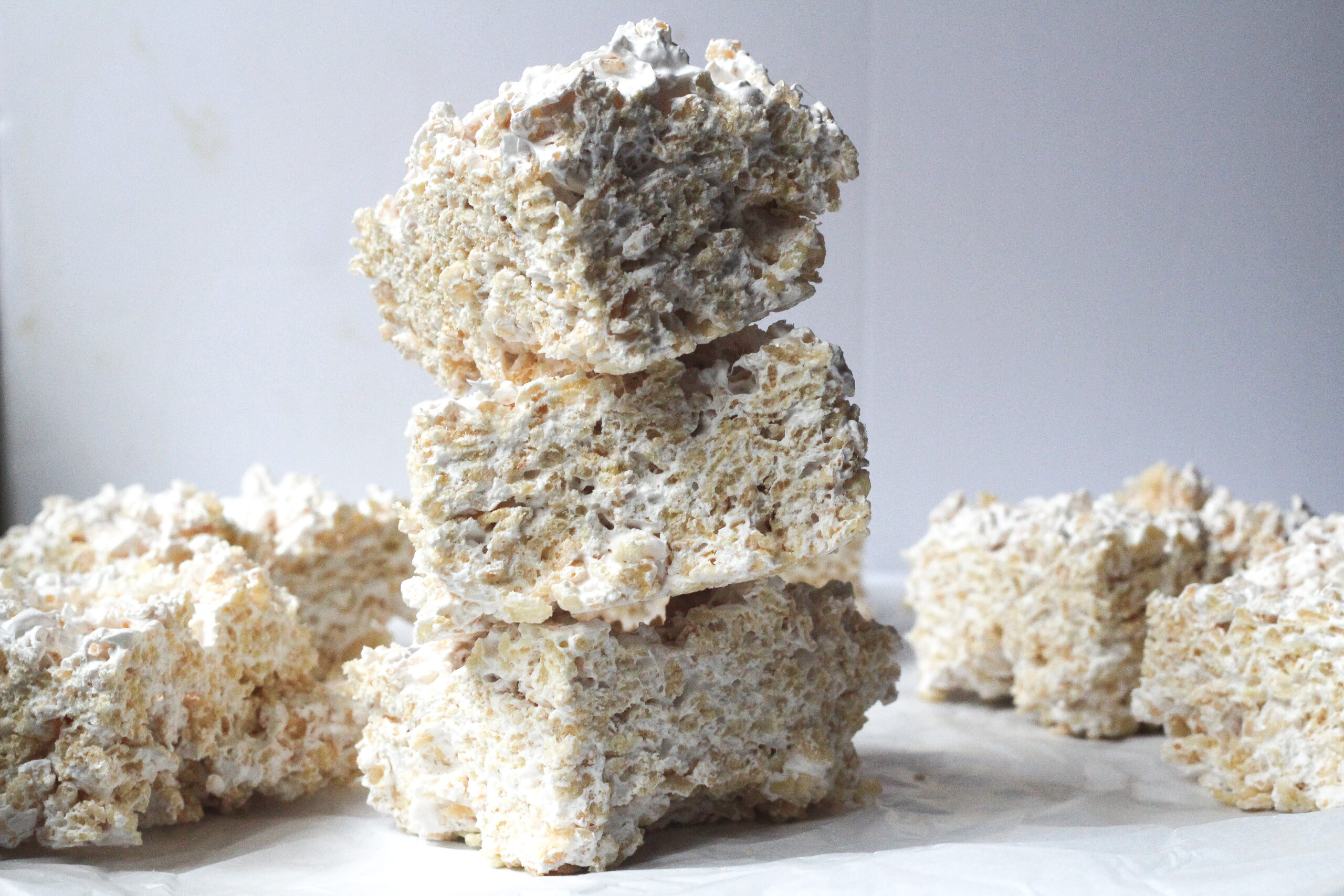 A stack of 3 maple rice krispie treats on a white marbled surface in front of a grey background and a few more maple rice krispie treats in a single layer