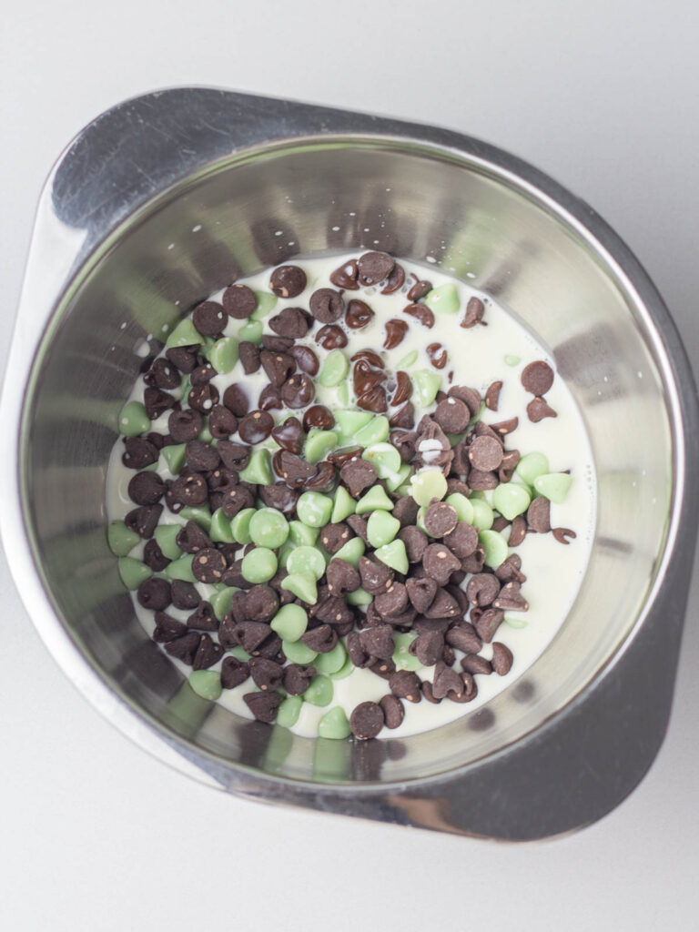 Warm heavy cream and mint chocolate chips in a small silver mixing bowl.