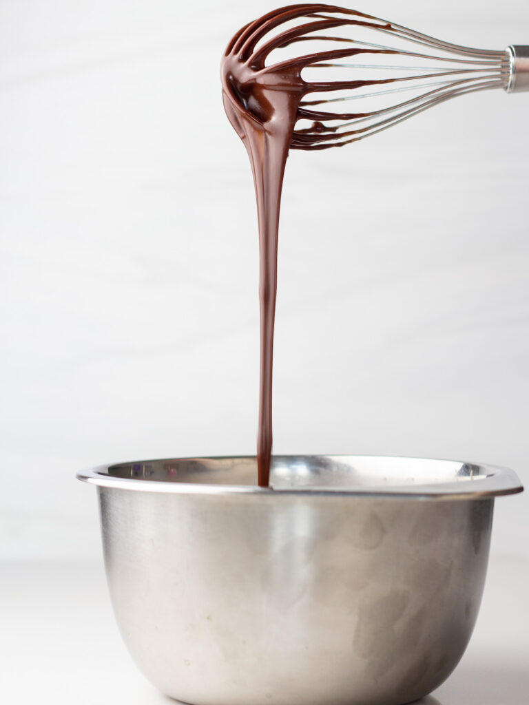 Smooth chocolate ganache drizzling from whisk into small silver mixing bowl.