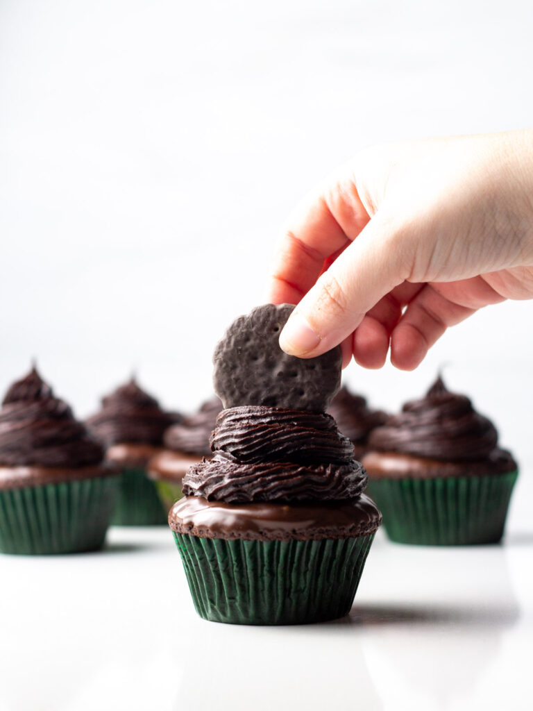 A hand placing a Thin Mint cookie on top of a frosted cupcake with more frosted cupcakes in the background.