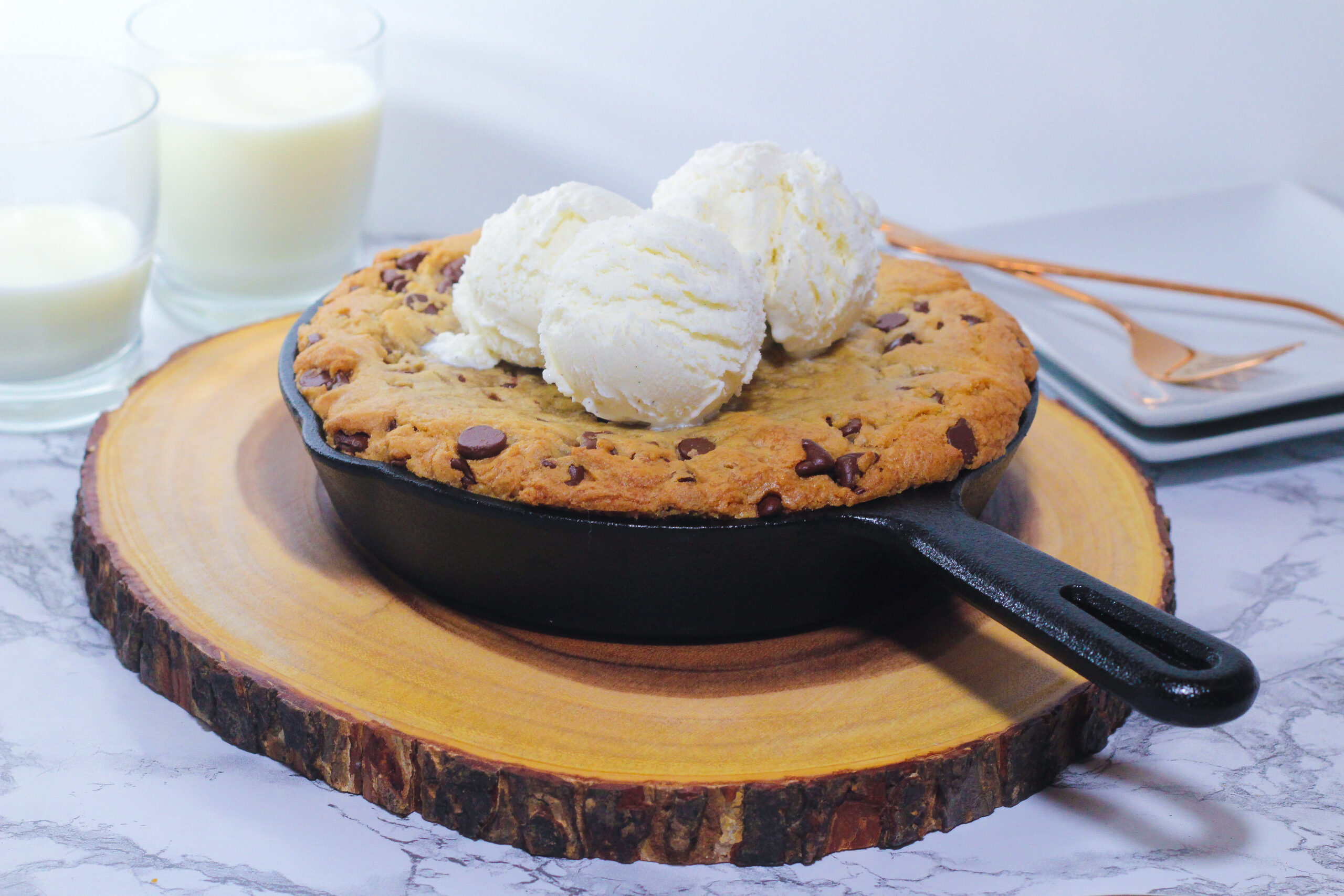 cast iron cookie topped with three scoops of vanilla ice cream in the skillet on a round piece of wood with two glasses of milk in the top left corner, one full and one less than half full, and two square white plates in the top right corner with two rose gold forks on them, all on a marbled surface