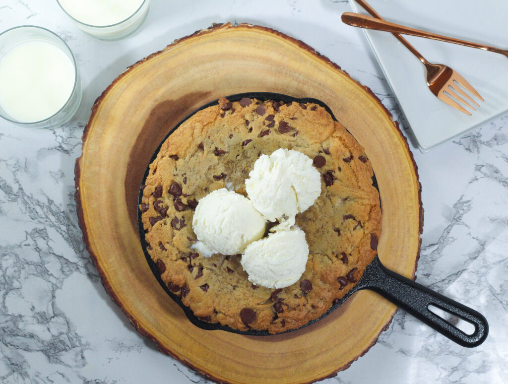 top down view of cast iron cookie topped with three scoops of vanilla ice cream in the skillet on a round piece of wood with two glasses of milk in the top left corner, one full and one less than half full, and two square white plates in the top right corner with two rose gold forks on them, all on a marbled surface