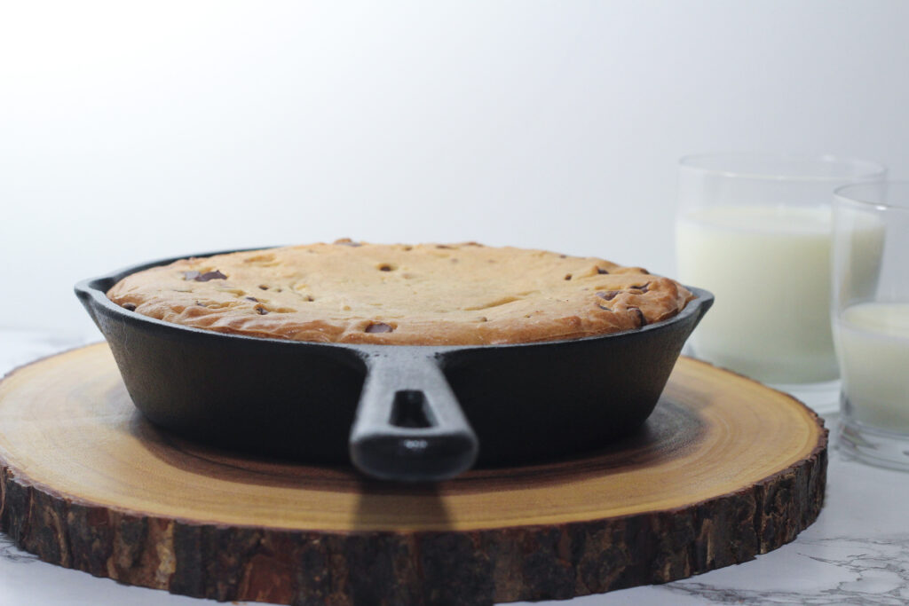 Side view of a cast iron cookie in a skillet looking on from the handle side of the skillet on a circular piece of wood with two glasses of milk partially out of the frame to the upper right, one glass full, one less than half full