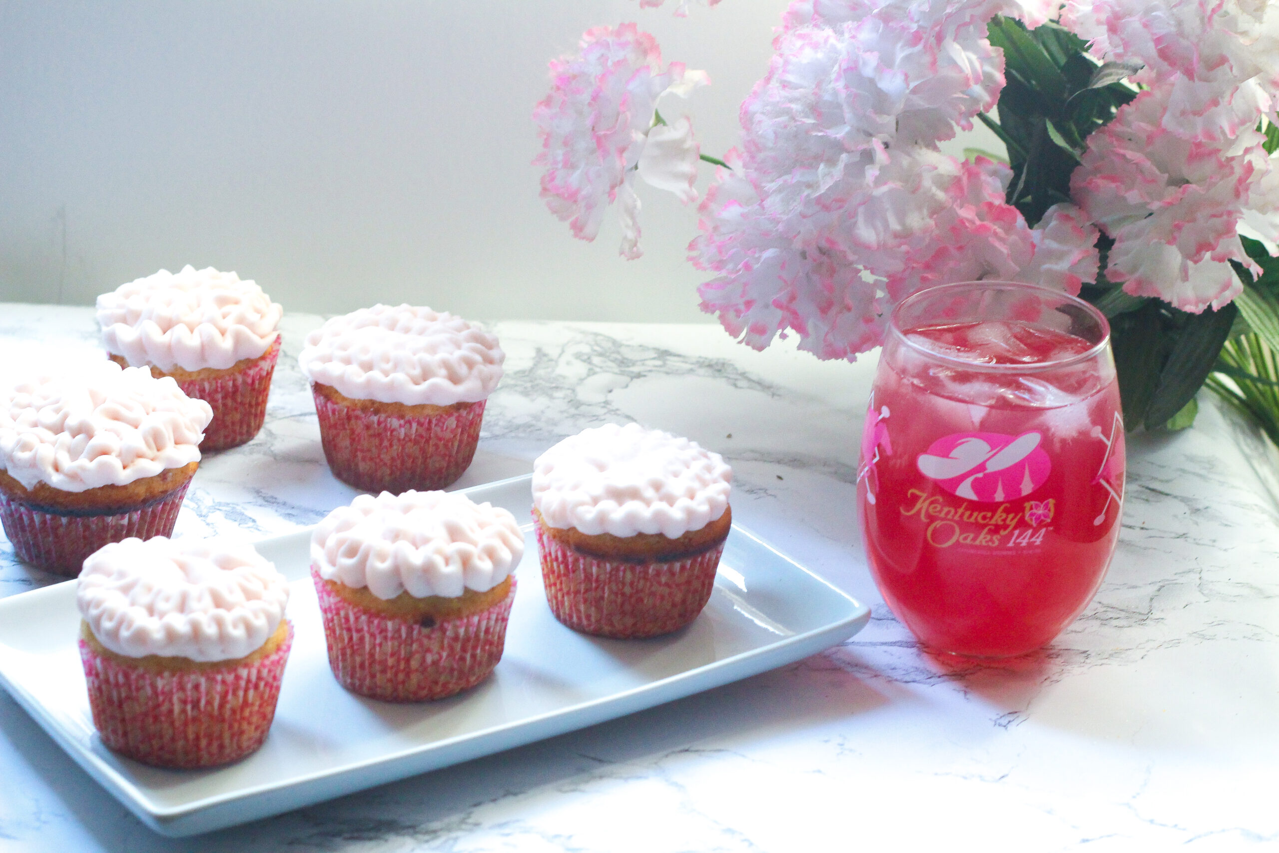 three lily cupcakes on a rectangle plate with more lily cupcakes behind the plate on the left side of the frame. An Oaks Lily in a stemless Oaks Day wineglass with pink and white flowers in the background