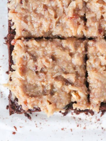 top down view of the corner of a batch of german chocolate brownies topped with a traditional coconut pecan frosting. The brownies are sliced into squares. The bottom left brownie is the only one fully in the shot, and the brownie above it, to the upper right, and to the right are all partially in frame.