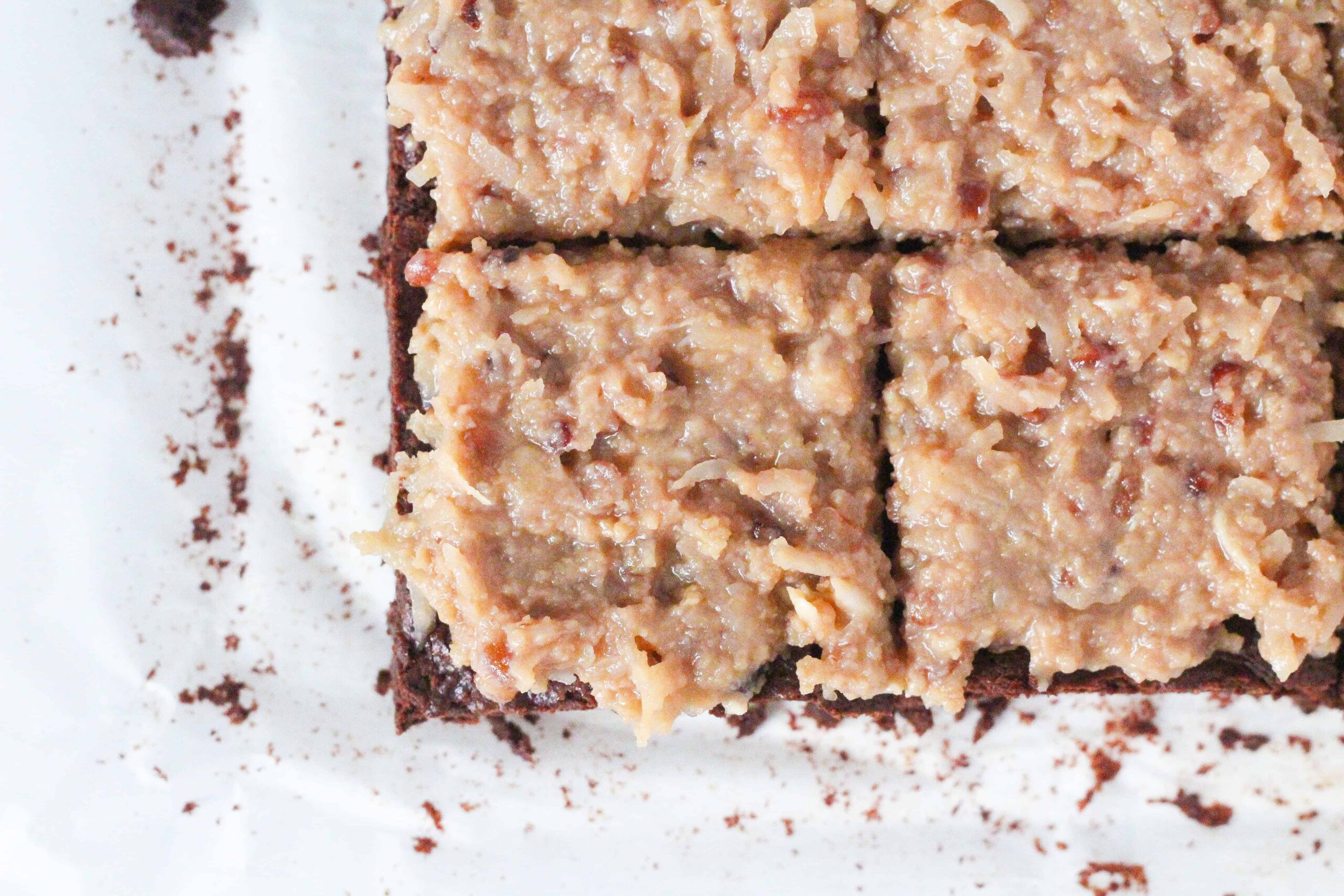 top down view of the corner of a batch of german chocolate brownies topped with a traditional coconut pecan frosting. The brownies are sliced into squares. The bottom left brownie is the only one fully in the shot, and the brownie above it, to the upper right, and to the right are all partially in frame.