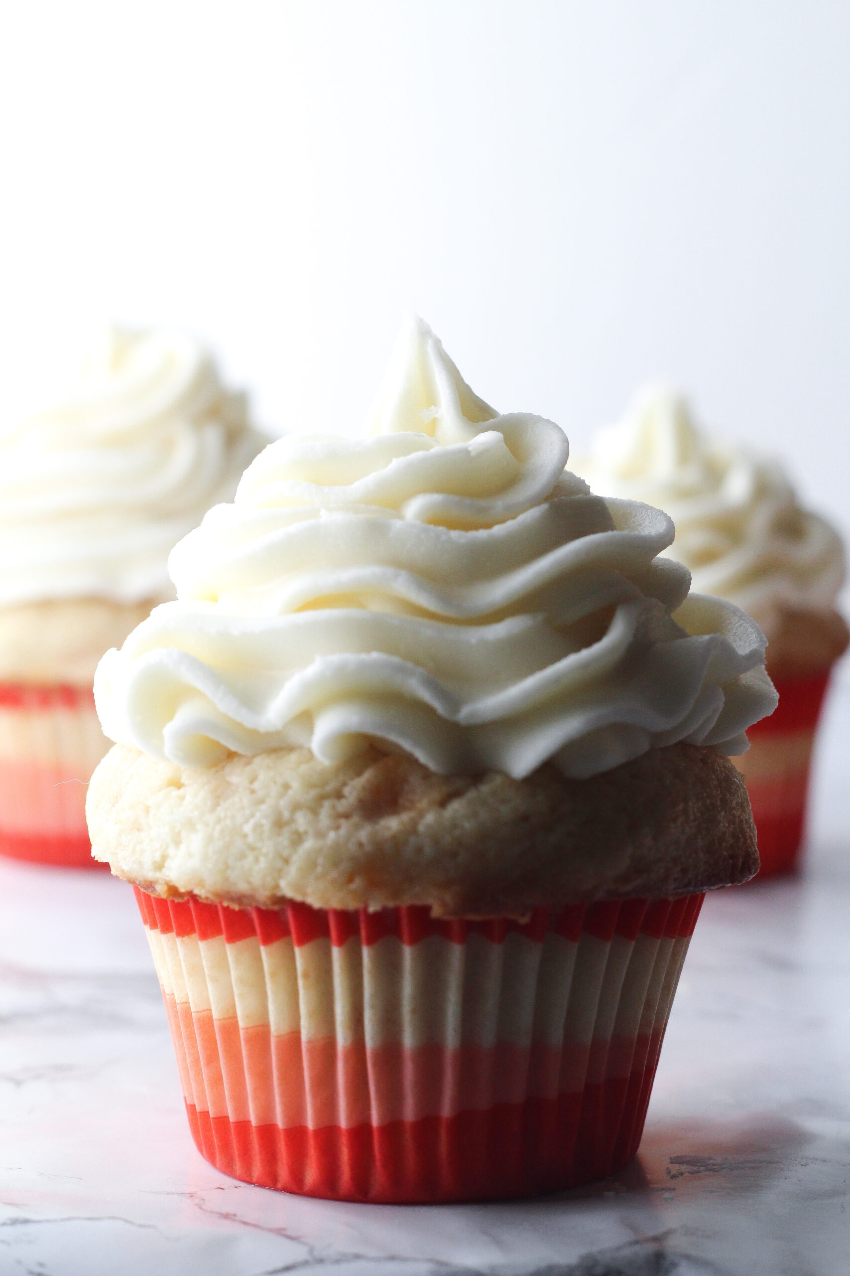 Close up of a bourbon peach sweet tea cupcake with two cupcakes in the back partially blocked by the one up front, all in red, white, and pink wrappers on a marbled surface in front of a white background