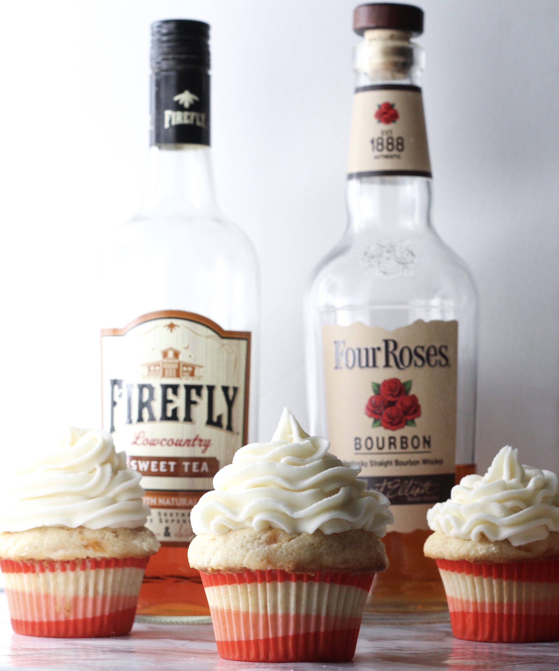 three bourbon peach sweet tea cupcakes in front of a bottle of firefly sweet tea vodka and a bottle of four roses bourbon