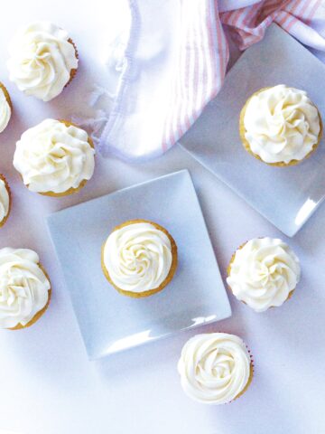 top down view of wannamango cupcakes on a white surface, each of two cupcakes are on square white plate. In the top right corner of the frame is a white and peach colored cloth napkin.