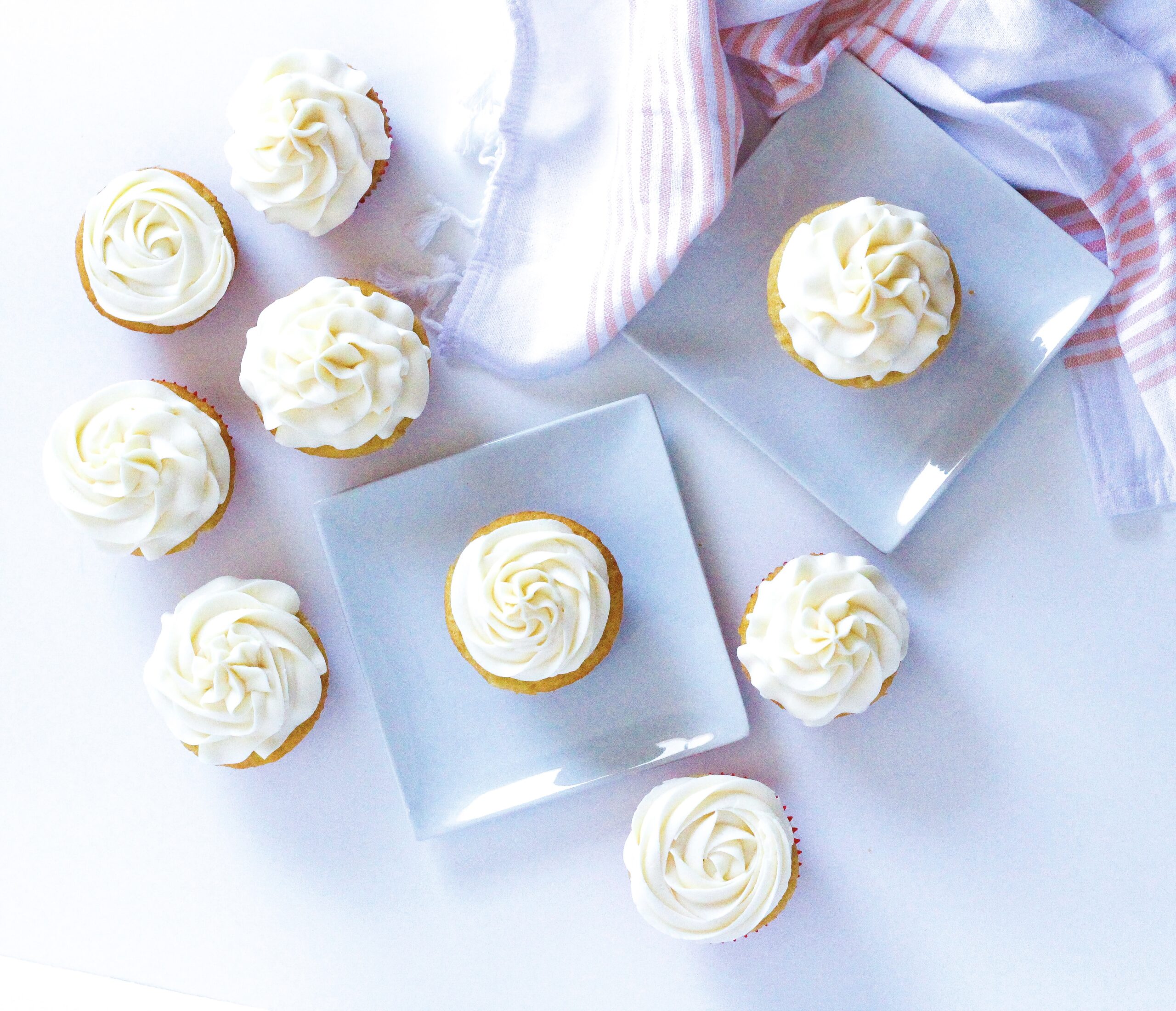 top down view of wannamango cupcakes on a white surface, each of two cupcakes are on square white plate. In the top right corner of the frame is a white and peach colored cloth napkin.