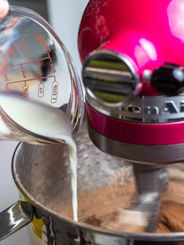 Measuring cup of milk being poured into a mixing bowl on a kitchen aid stand mixer.