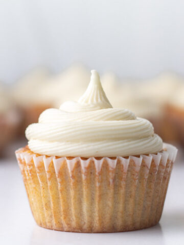 A Cinnamon Cupcake with marshmallow frosting close up front and center in the image with a row of cupcakes blurred in the background.