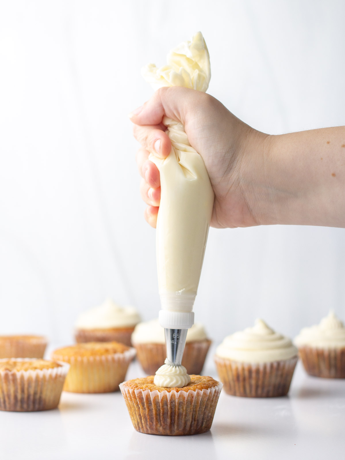 A hand piping marshmallow frosting onto a cinnamon cupcake with frosted and unfrosted cupcakes in the background.