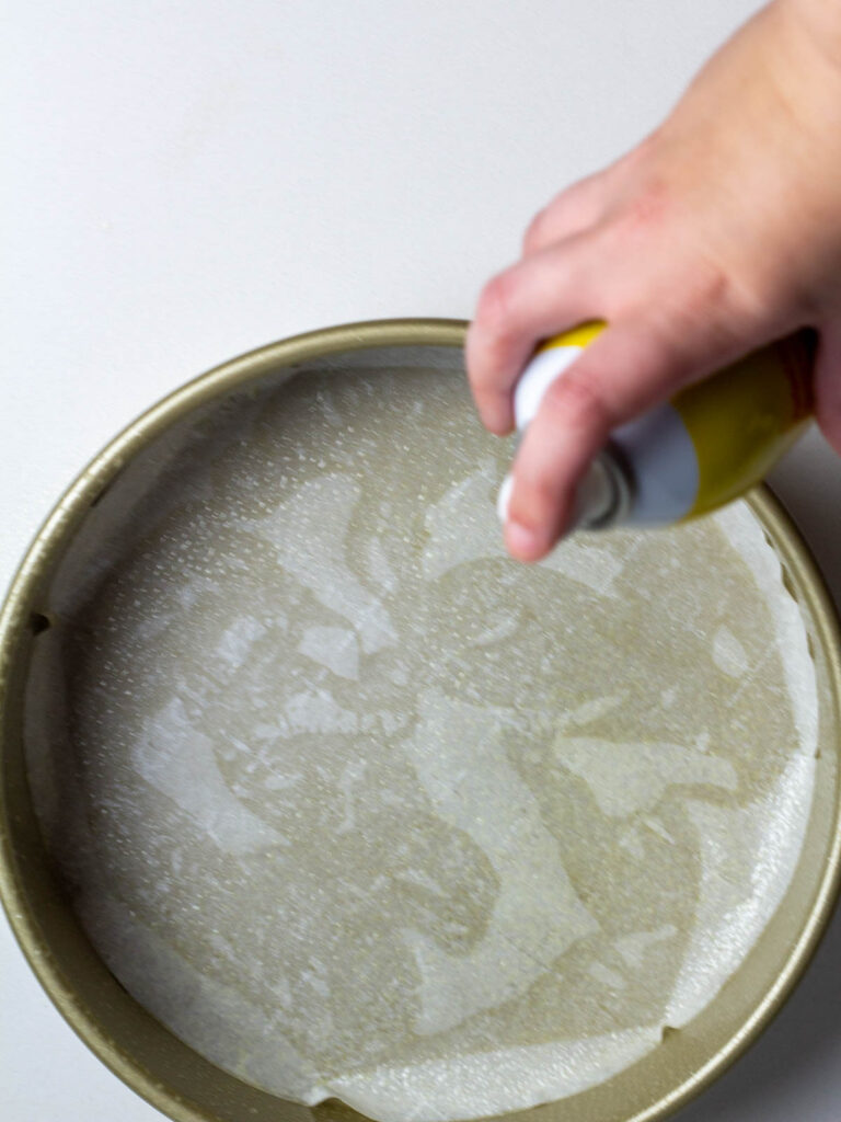 A hand spraying non-stick spray into a parchment lined cake pan.