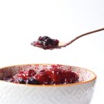 a rose gold spoon full of cranberry sauce being held above a white textured bowl with an orange rim full of cranberry sauce