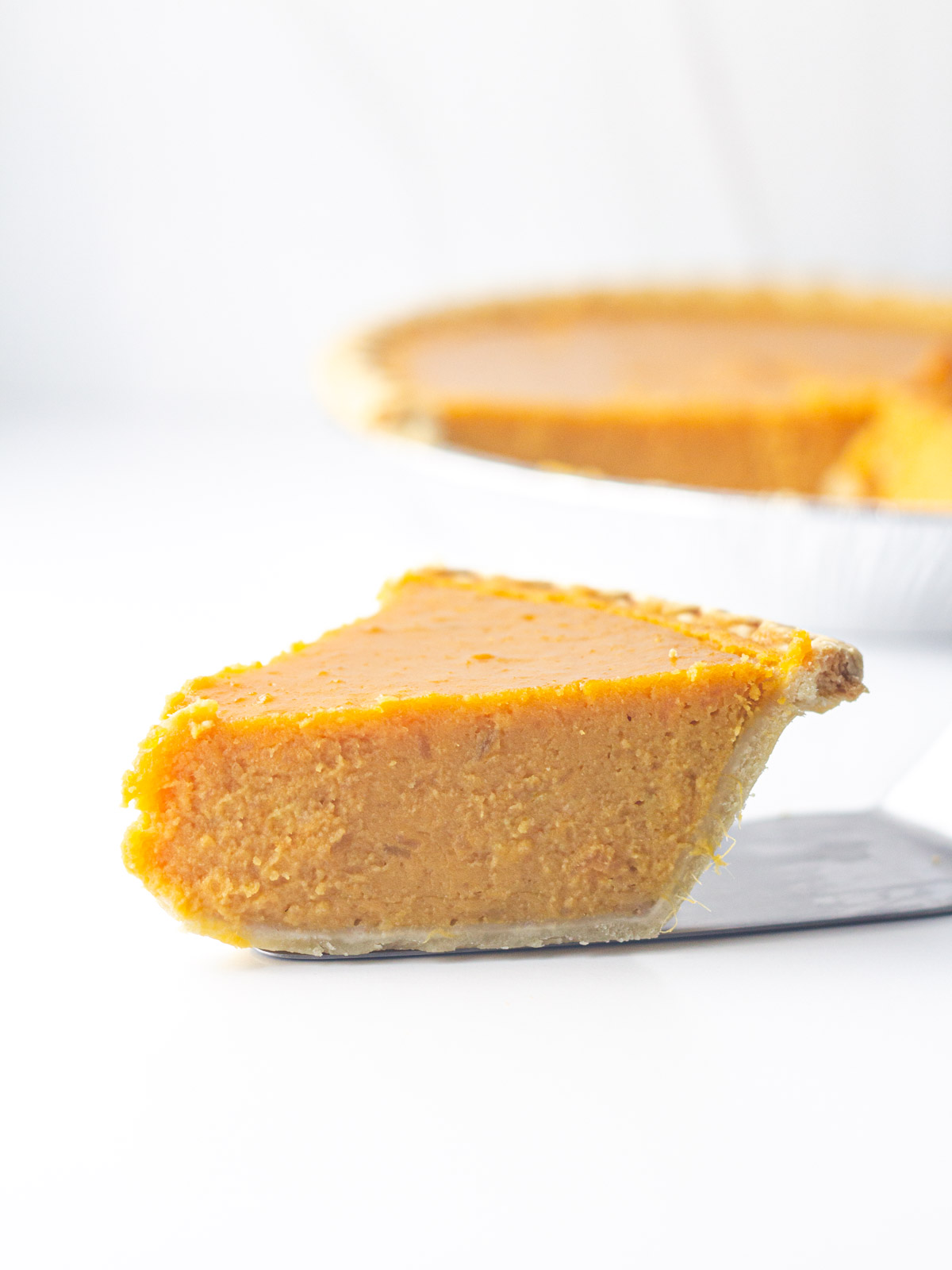A slice of sweet potato pie on the pie slicer on a white surface in front of the rest of the pie blurred in the background going out of frame to the right.