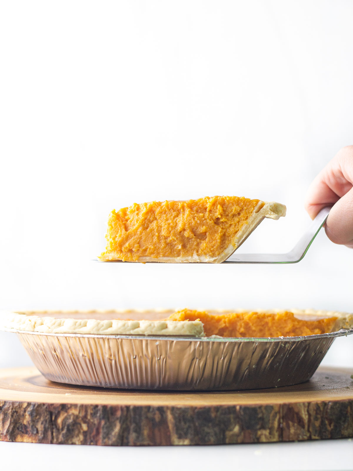 A slice of sweet potato pie being lifted out of the rest of the pie dish which is sitting on a round wooden plank.