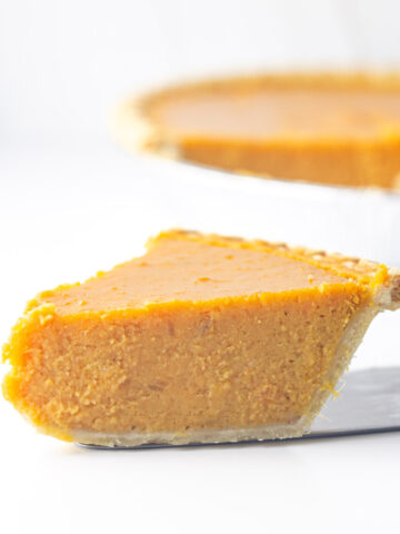 A slice of sweet potato pie on the pie slicer on a white surface in front of the rest of the pie blurred in the background going out of frame to the right.