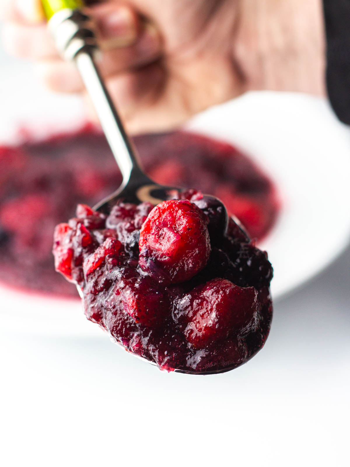 A hand holding a spoon full of bourbon cranberry sauce close to the camera with the bowl of the rest of the cranberry sauce blurred in the background.