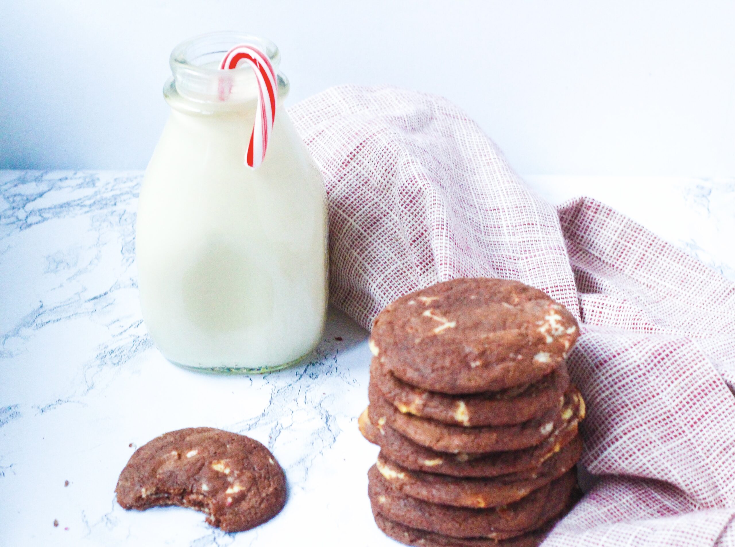 In the front right of the image is a stack of fudgy peppermint chocolate cookies. To the right of the cookie stack is a red and white napkin that runs toward the back left of the frame where there is a glass jar of milk with a candy cane in it. In front of the milk jar and to the left of the cookie stack is a fudgy peppermint chocolate cookie with a bite taken out of it.