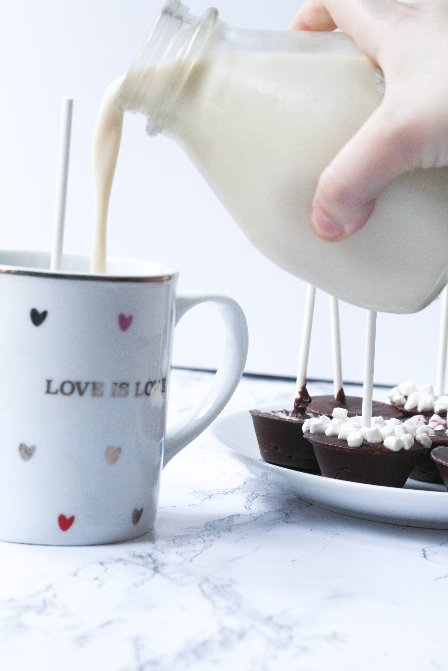 A white mug covered in multi-colored hearts that says Love is Love with a stick coming out of the top of the mug (the Nutella hot chocolate on a stick is in the mug, but you can't actually see the chocolate). coming in from the right side of the frame is a hand holding a glass jar of milk and pouring the milk into the mug. Beneath the jar on the right side of the frame is a plate of nutella hot chocolates on sticks, with the plate cut off on the right side of the frame. The most visible hot chocolate stick has mini marshmallows covering the top of the chocolate.