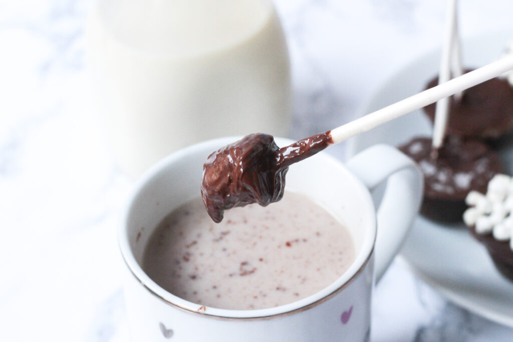 a mostly melted nutella hot chocolate on a stick hovering over a white mug with a gold lip with hot chocolate in side. Behind the mug is a cut off glass jar of milk.