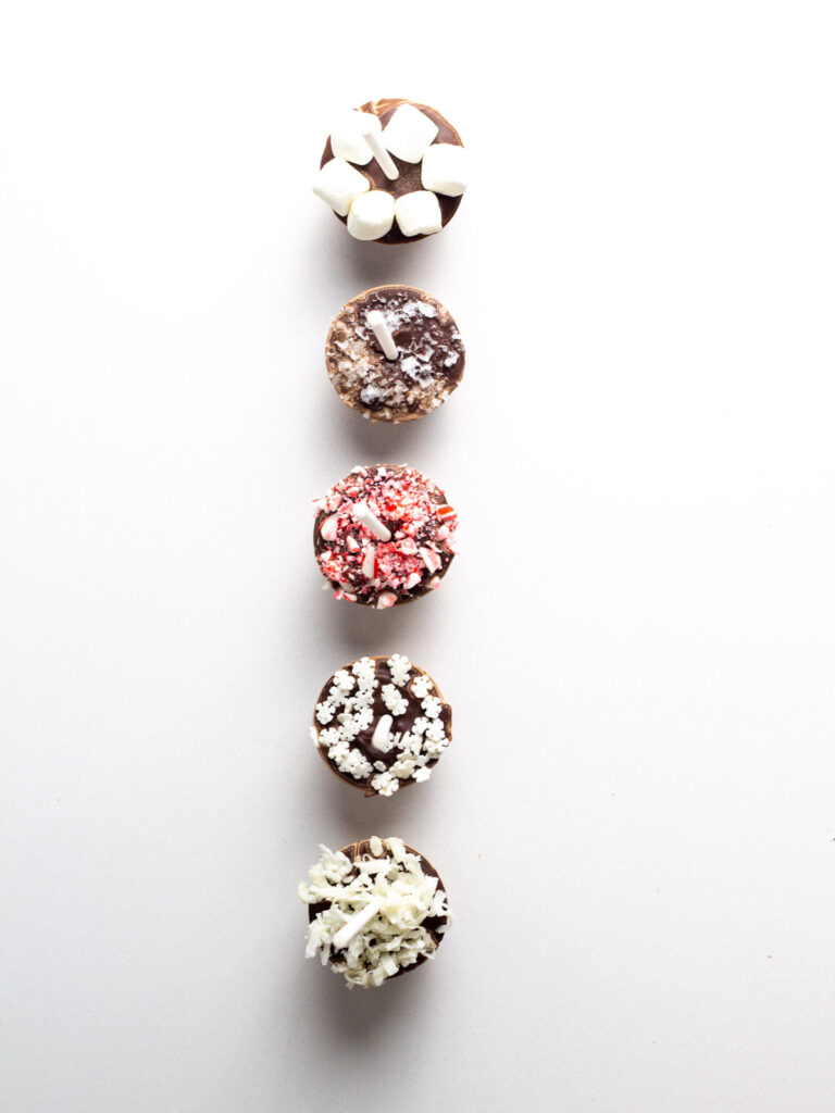 A top down view of a row of 5 hot chocolates on sticks with various toppings.