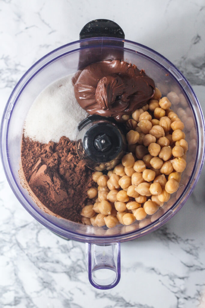 overhead shot of the ingredients in the food processor, the chickpeas toward the bottom right, the Nutella top center, the sugar top left, and the cocoa powder is bottom left, all on top of a marble surface