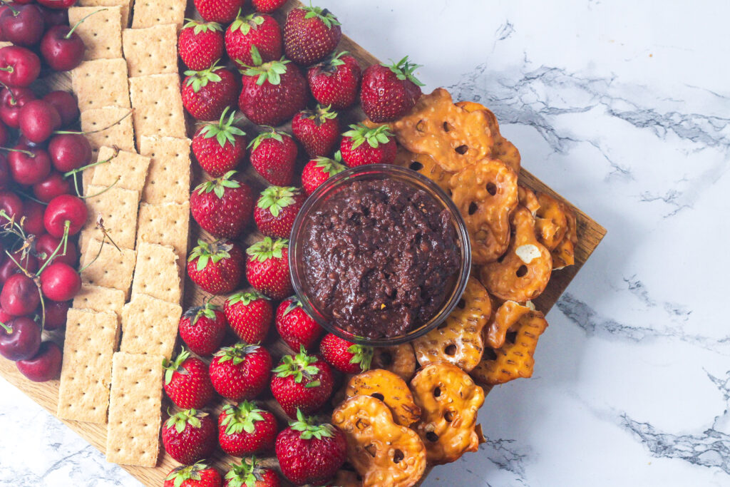 Nutella Hummus in a clear bowl on a tray with rows, from left to right, of cherries, graham crackers, strawberries, and pretzel crisps. The cherries are out of frame in the top left of the image and the board comes into the frame at an angle
