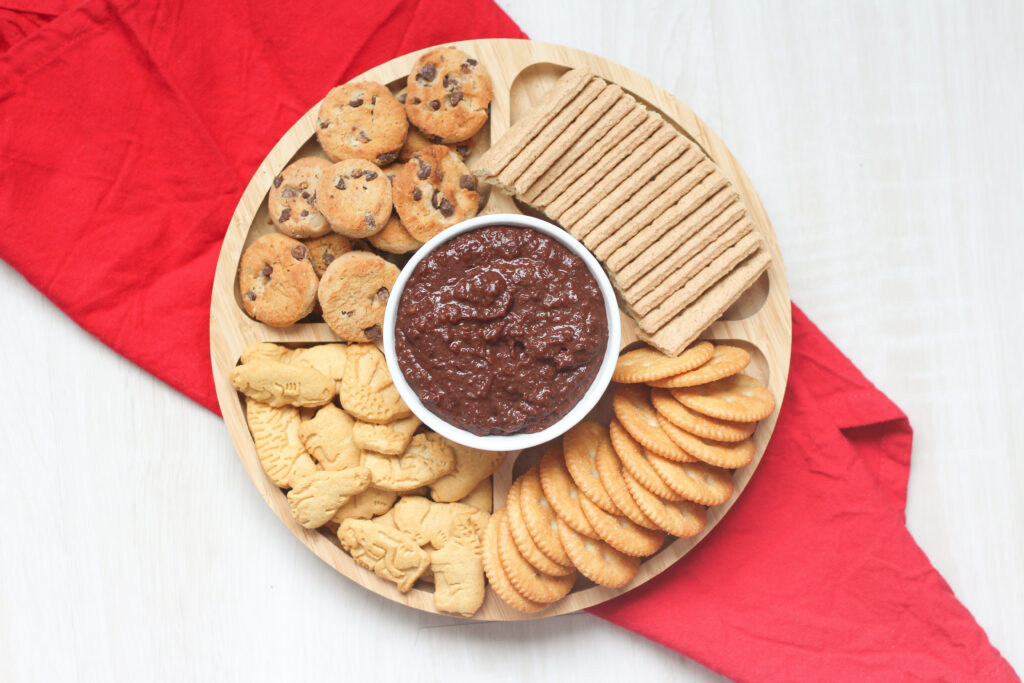 tray of cookies and crackers around a white bowl of Nutella hummus on a red linen.