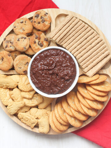 tray of cookies and crackers around a white bowl of Nutella hummus on a red linen.