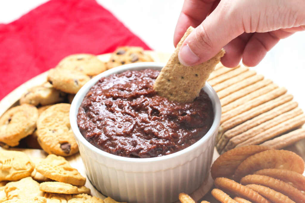 a hand dipping a graham cracker into a white bowl of Nutella hummus.