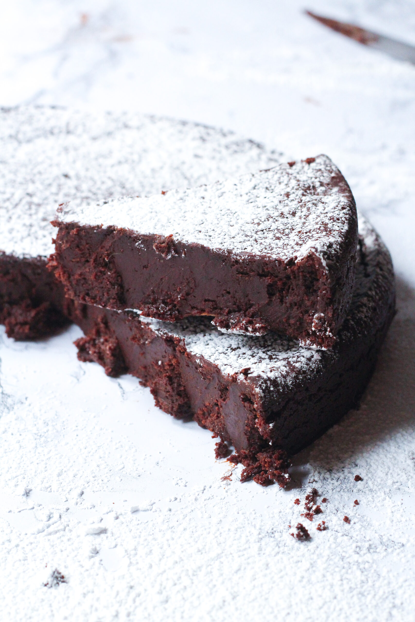 a slice of red wine flourless chocolate cake sitting on top of the remainder of the remainder of the cake from which the slice was removed, but slightly angled so that it doesn't line up exactly with the cake. The cake and the slice are dusted with powdered sugar.