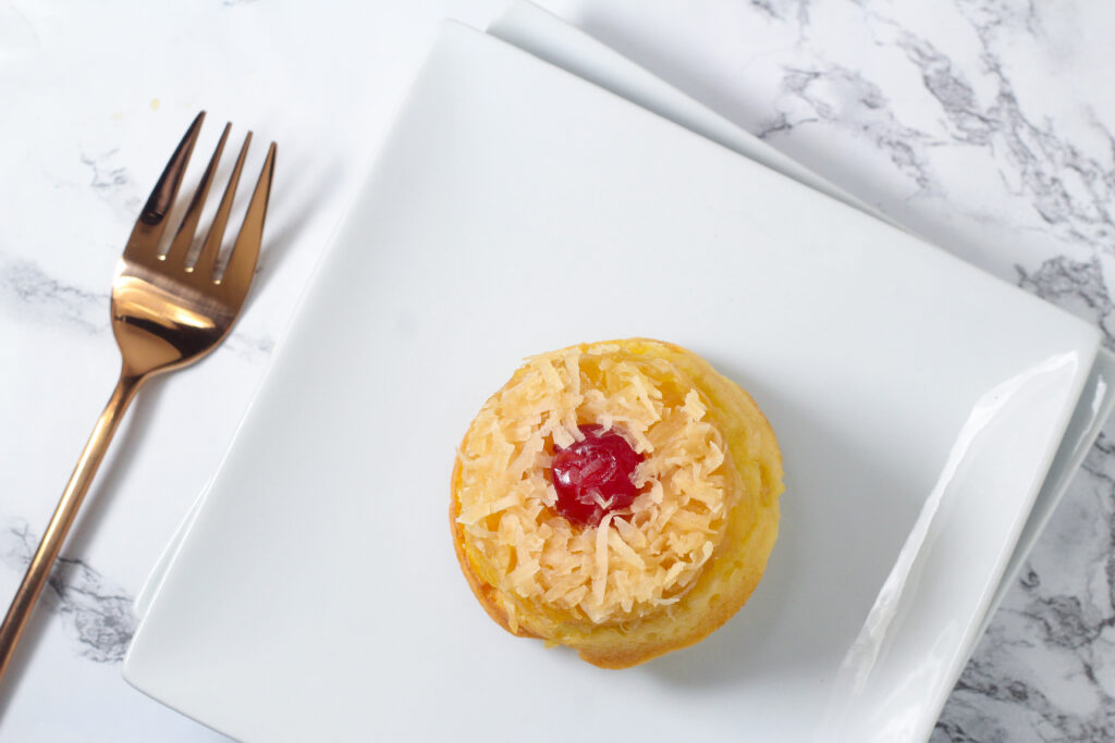 top down view of a single piña colada upside down cupcake on a square white plate stacked on top of another square white plate with a rose gold fork to the left of the plates all on a marbled surface