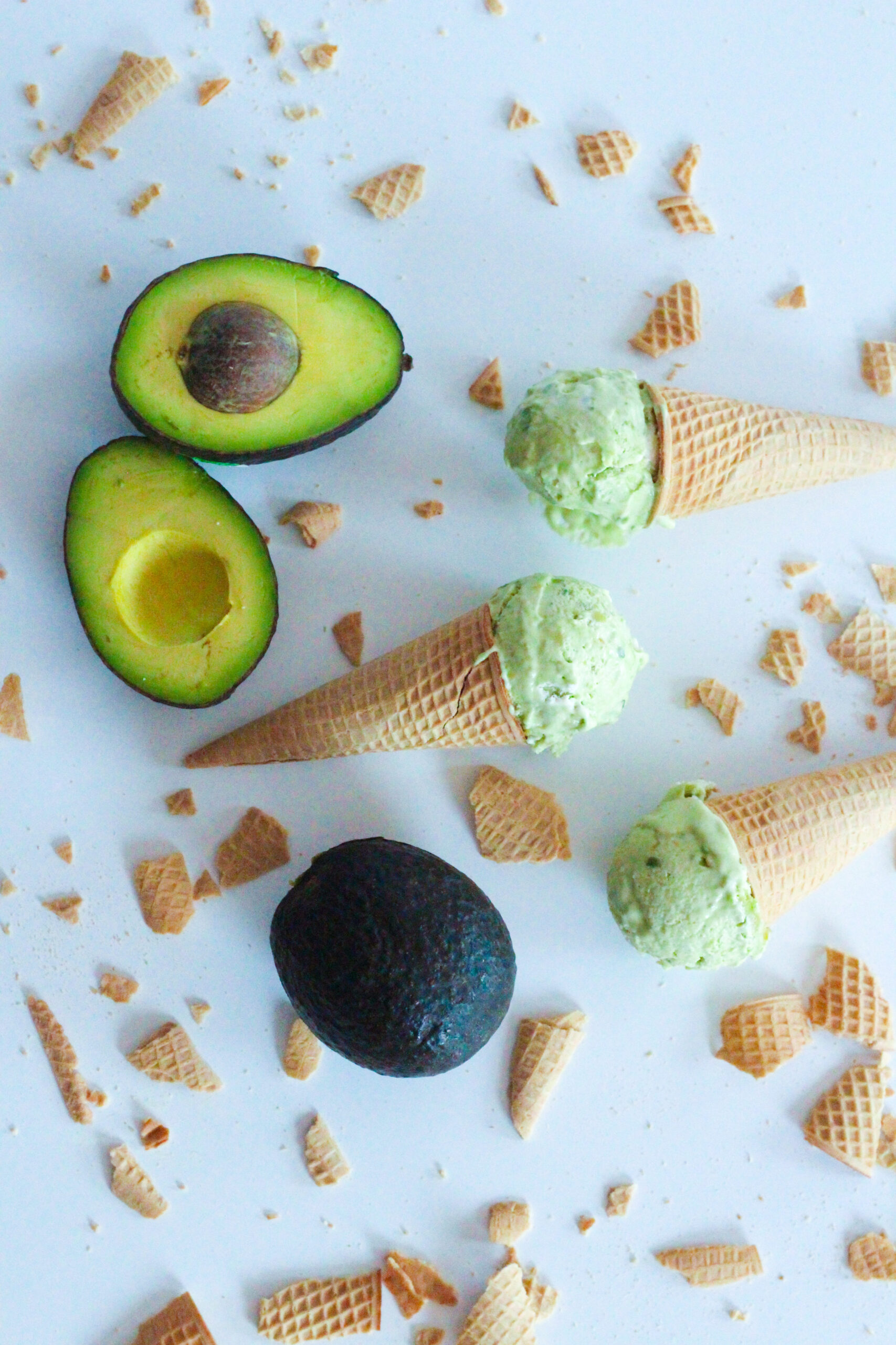 Top down view of three cones, each with one scoop of avocado ice cream on each cone. The cones are laying on their sides on a white surface, facing alternate directions. There are two halves of an avocado in the upper left corner and a whole avocado in the lower center. There are broken pieces of ice cream cones surrounding the cones and avocados.