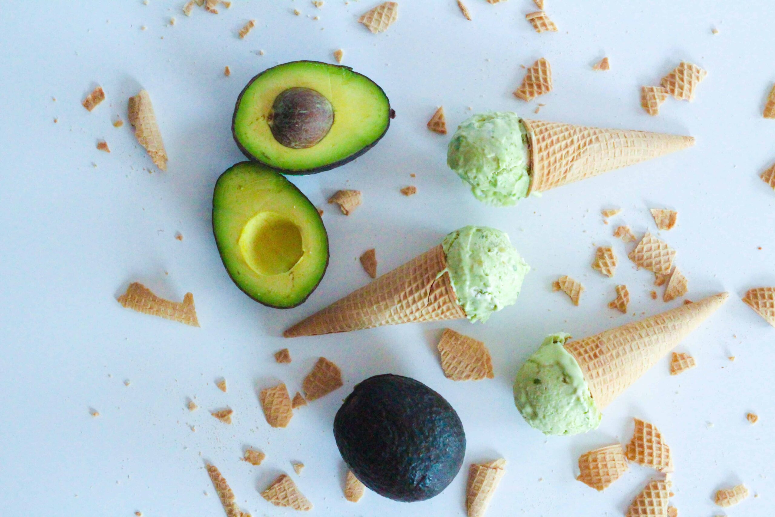 Top down view of three cones, each with one scoop of avocado ice cream on each cone. The cones are laying on their sides on a white surface, facing alternate directions. There are two halves of an avocado in the upper left corner and a whole avocado in the lower center. There are broken pieces of ice cream cones surrounding the cones and avocados.
