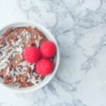 top down view of a white textured ramekin of chocolate grits topped with coconut shavings and three raspberries, sitting on a marbled surface