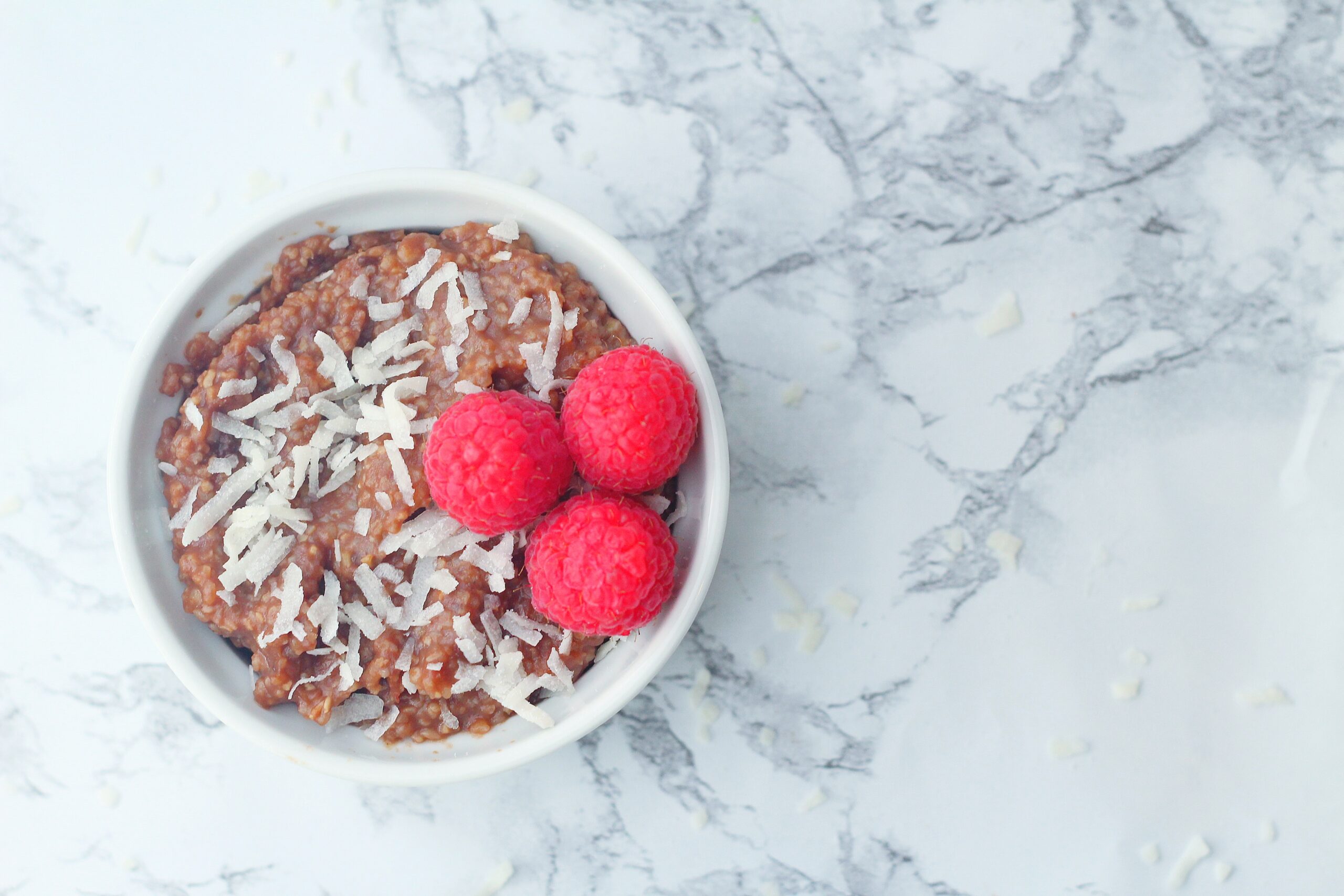 top down view of a white textured ramekin of chocolate grits topped with coconut shavings and three raspberries, sitting on a marbled surface