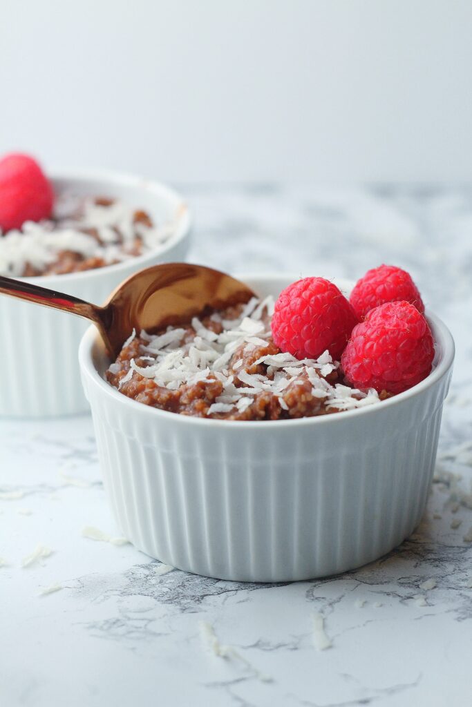 white textured ramekin of chocolate grits topped with coconut shavings and three raspberries with a rose gold spoon in the grits, sitting on a marbled surface, with a partial view of a slightly blurred second ramekin of chocolate grits in the background