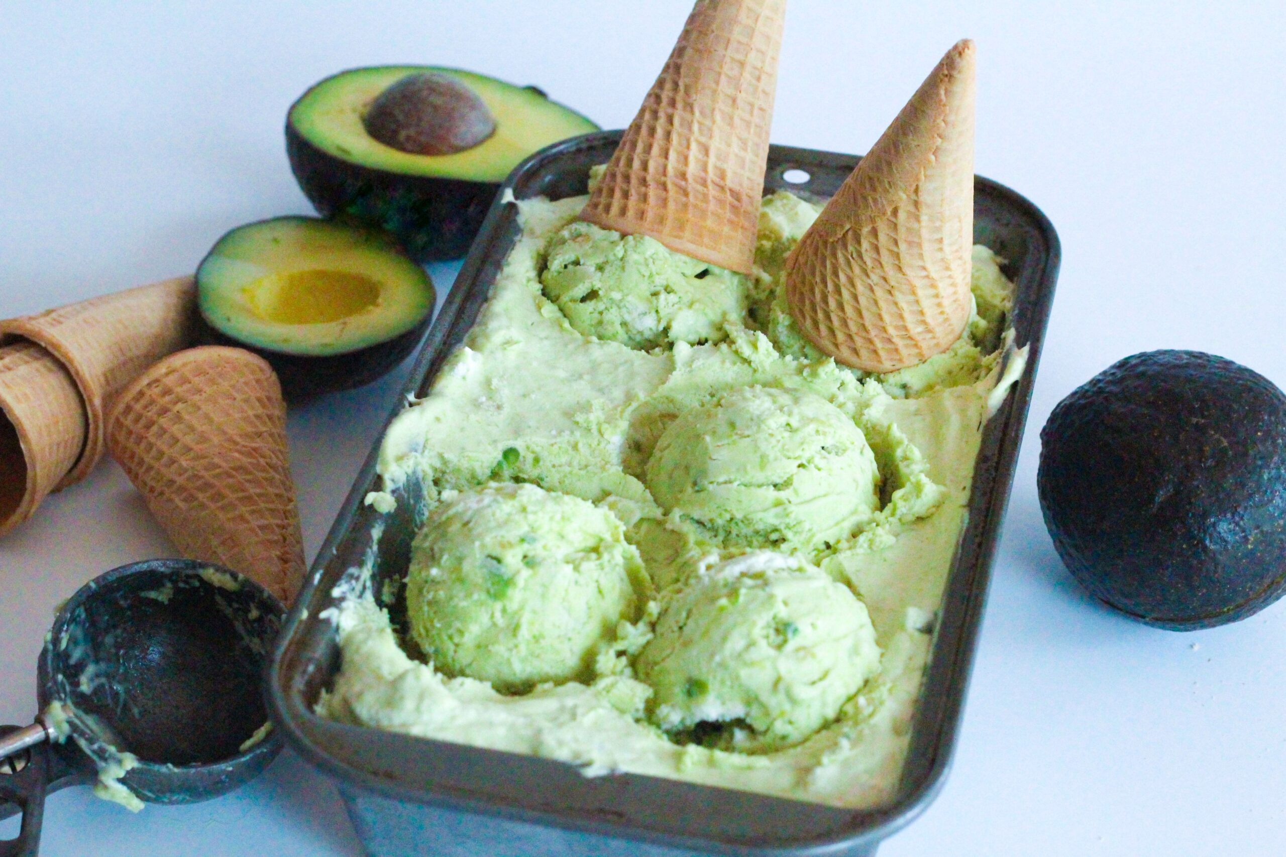 Angled view of the loaf pan of avocado ice cream with three scoops sitting in the pan, and two more with ice cream cones on them also still sitting in the pan. to the right of the pan is a whole avocado. To the left of the pan, on the upper side of the frame is two halves of an avocado, in front of them are three ice cream cones, two stacked inside of eachother, and in front of those is an ice cream scooper.